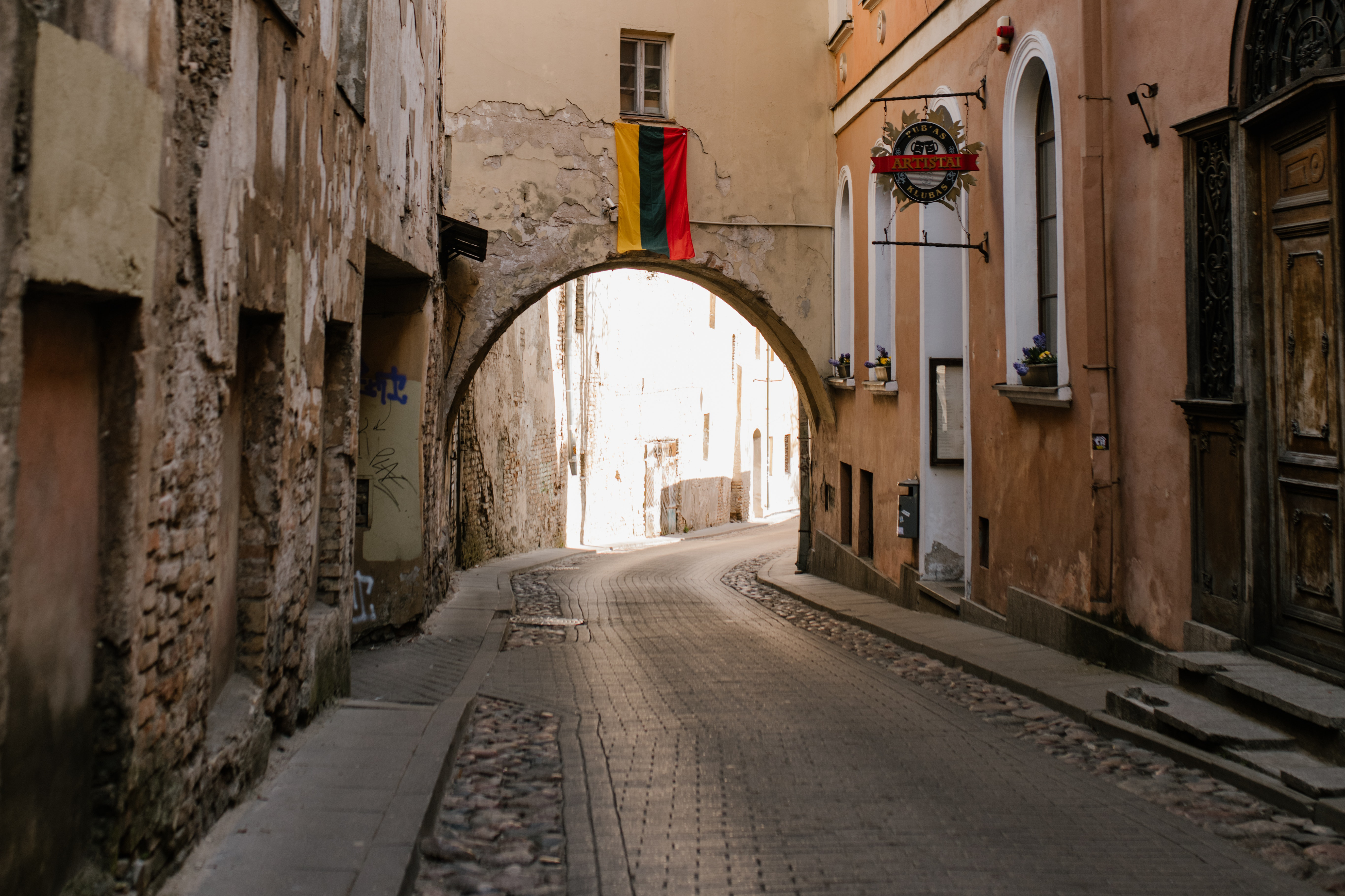 A cobblestone path. At the end of the path is an archway through which you can see the blue sky.
