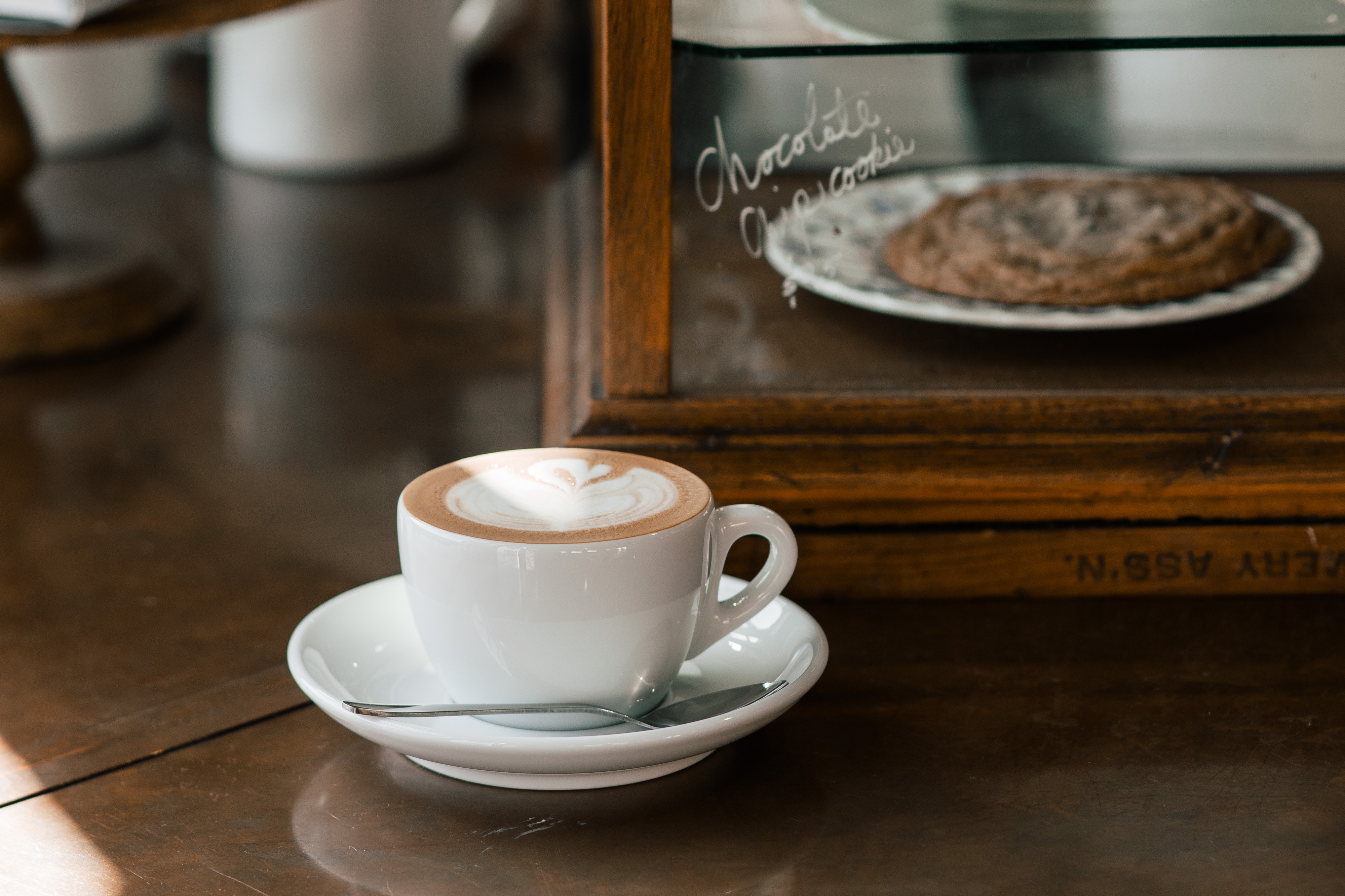 A steaming coffee sits on the counter in a ray of light, in front of a glass and wood case of deserts.